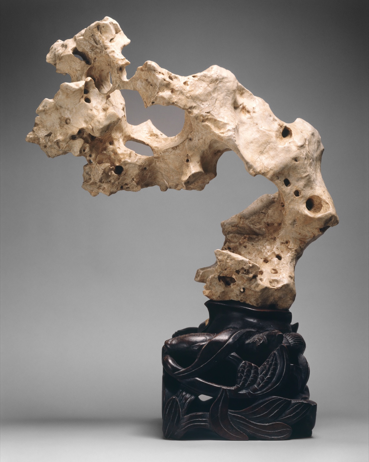 Une pierre calcaire blanche, creusée de trous et de cavités par l'eau, dressée sur un socle de bois sombre sculpté