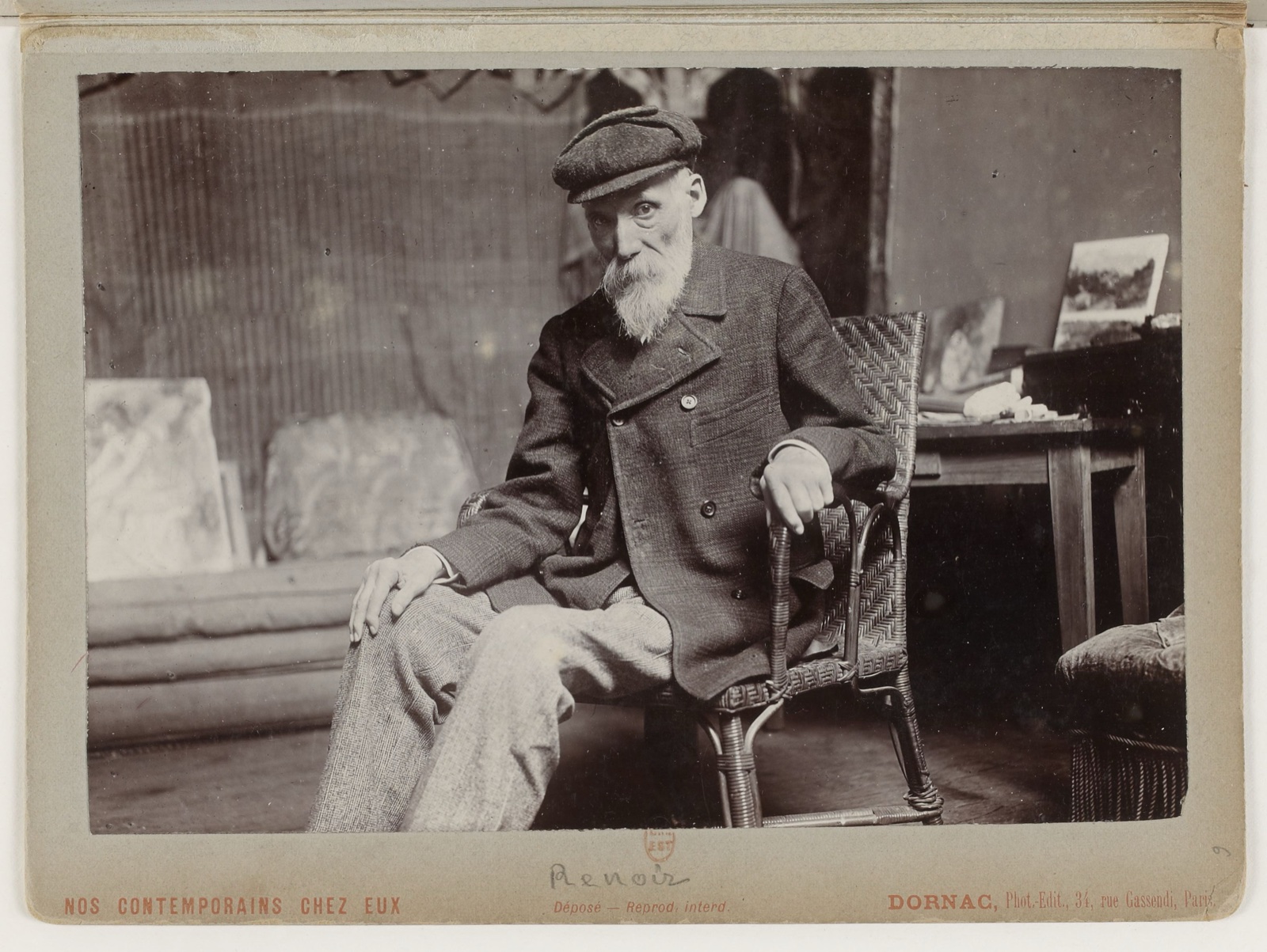 Photographie ancienne montée sur carton : un vieil homme à barbe blanche et casquette, assis dans un fauteuil d'osier, au milieu de toiles et de meubles d'atelier ; en bas, la mention imprimée « Nos contemporains chez eux »