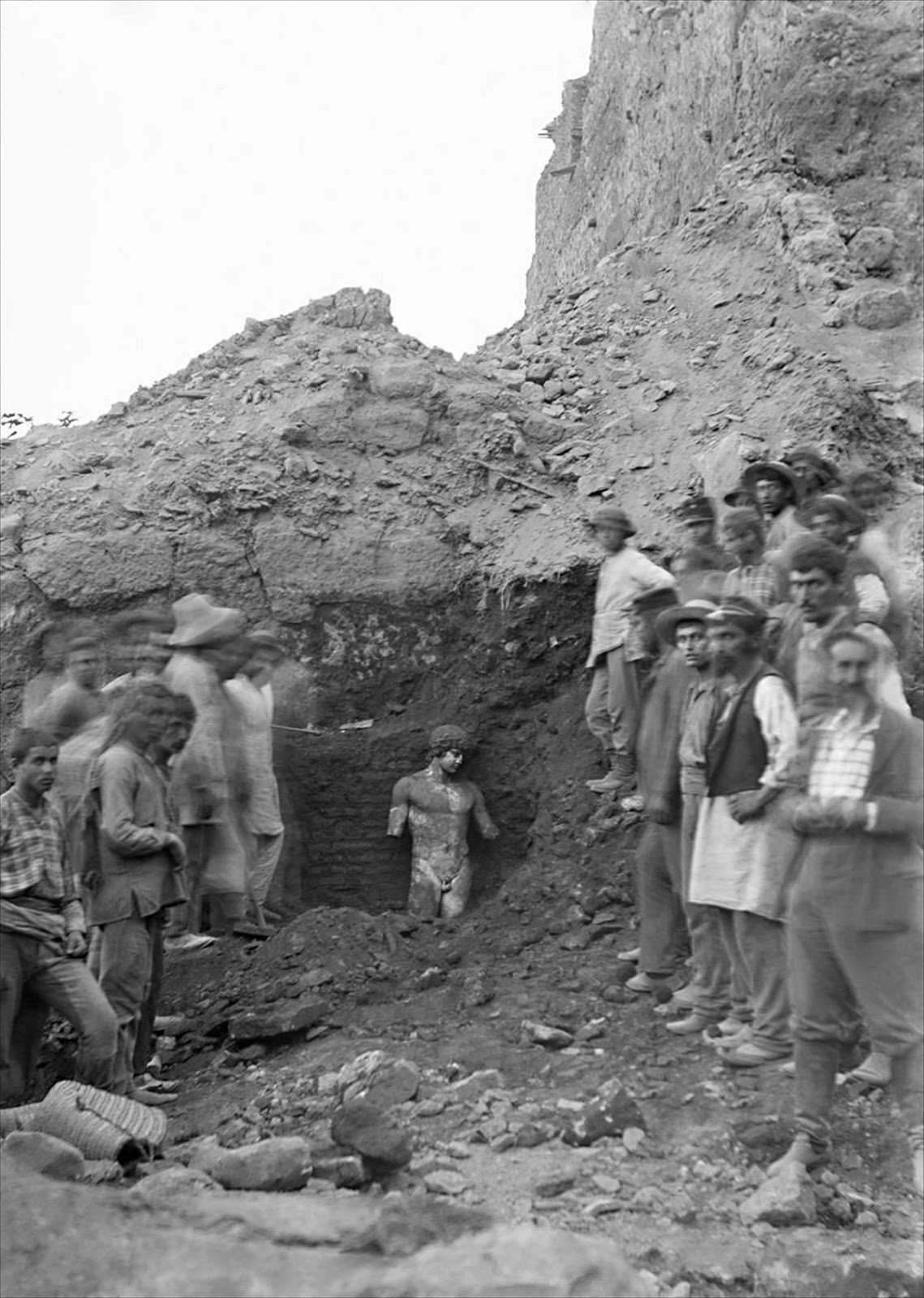 Photographie ancienne : au fond d'une tranchée de fouille, une statue de marbre dégagée jusqu'à la taille, entourée d'ouvriers qui la regardent