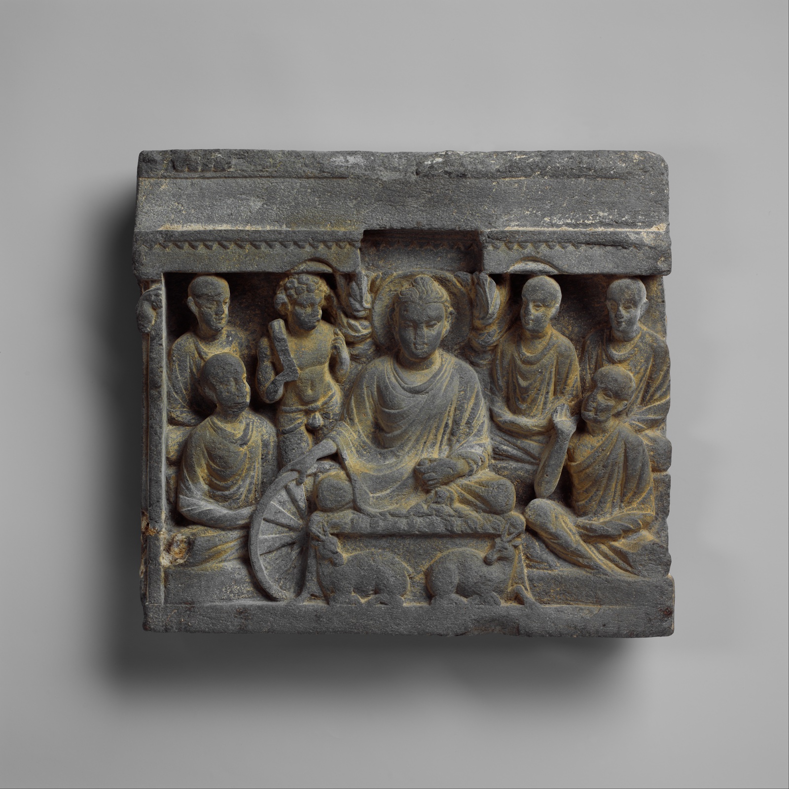 Relief de pierre grise : le Bouddha assis au centre, entouré de moines et de personnages ; à ses pieds, une roue et deux cerfs