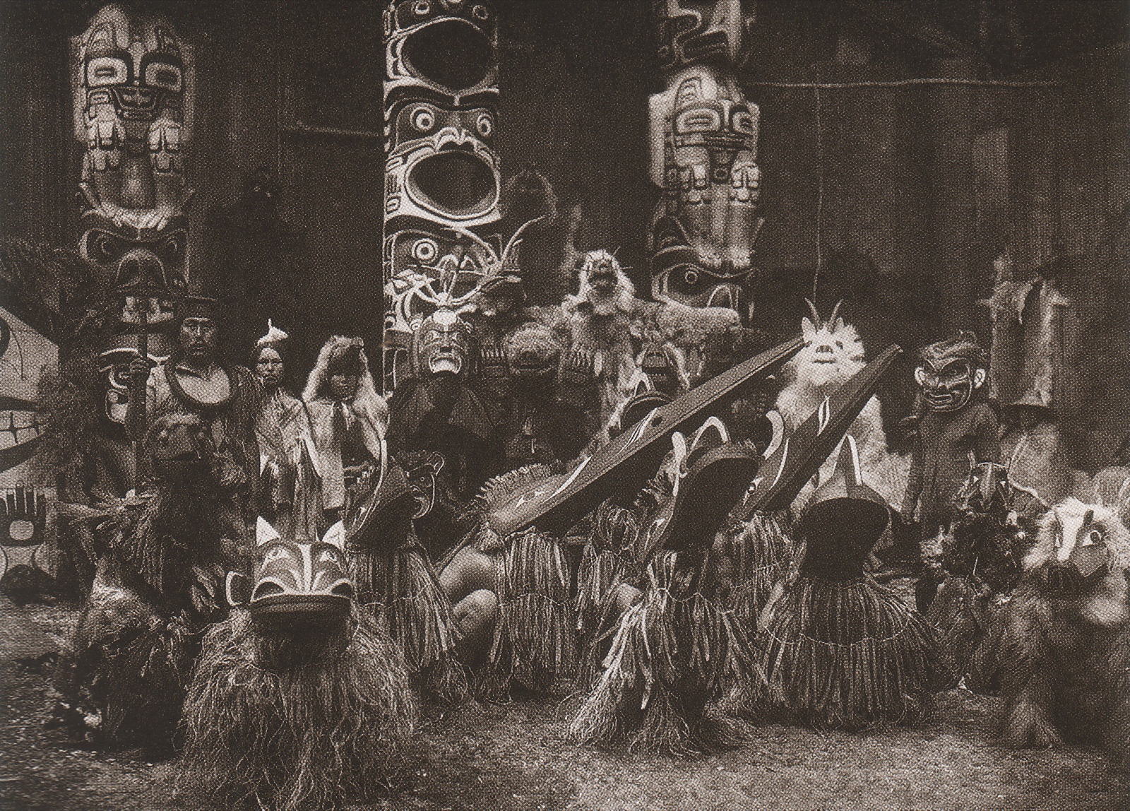 Photographie ancienne : devant de grands mâts sculptés, une troupe de danseurs vêtus de franges d'écorce porte des masques d'animaux à longs becs