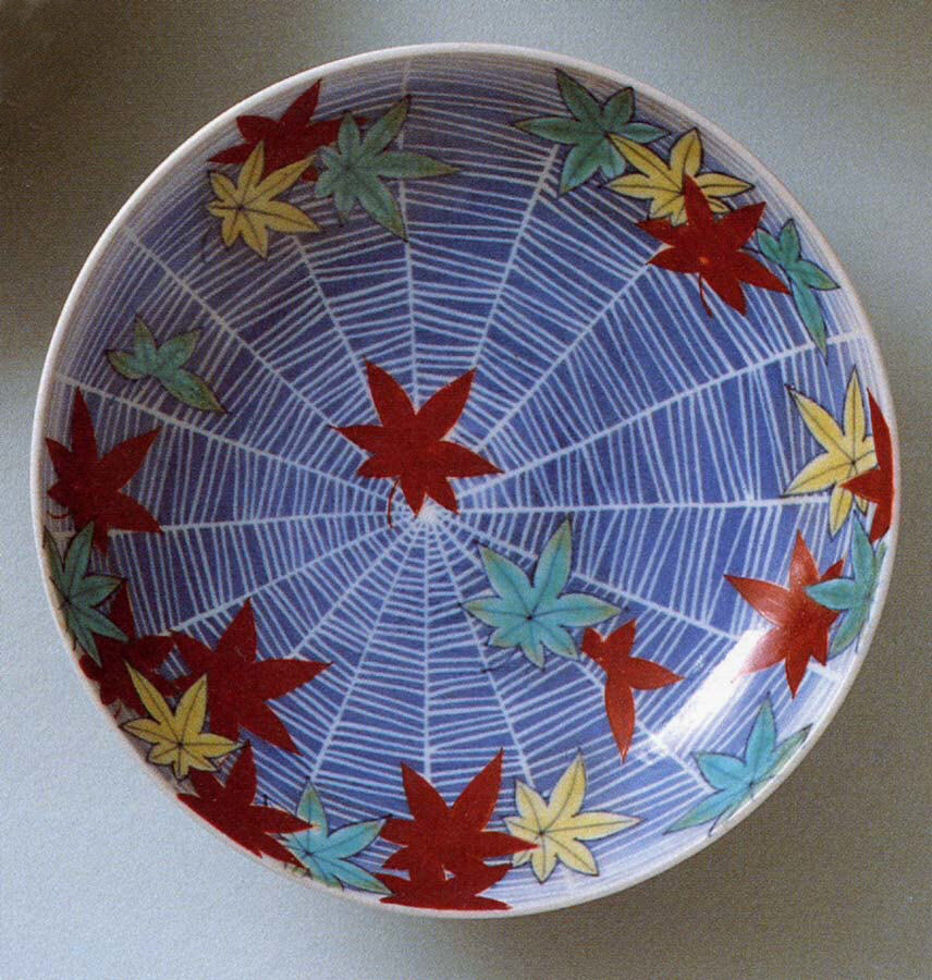 Coupelle de porcelaine au fond bleu parcouru d'une toile d'araignée blanche, sur laquelle tombent des feuilles d'érable rouges, jaunes et vertes