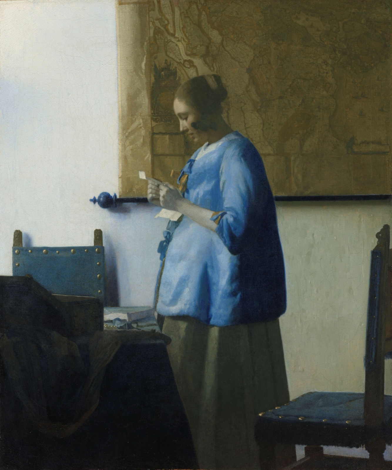 Une jeune femme en veste bleue, debout près d'une table, lit attentivement une lettre qu'elle tient à deux mains ; derrière elle, une carte est accrochée au mur