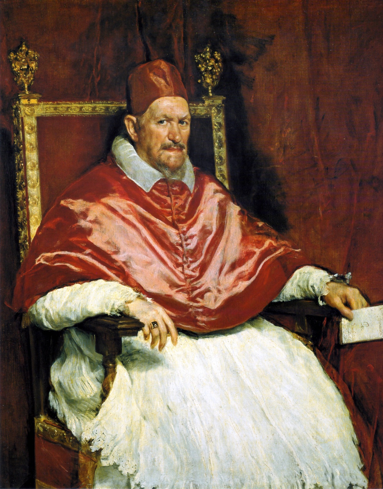 Le pape assis dans un fauteuil doré, en camail et barrette rouges, le regard dur, une lettre à la main