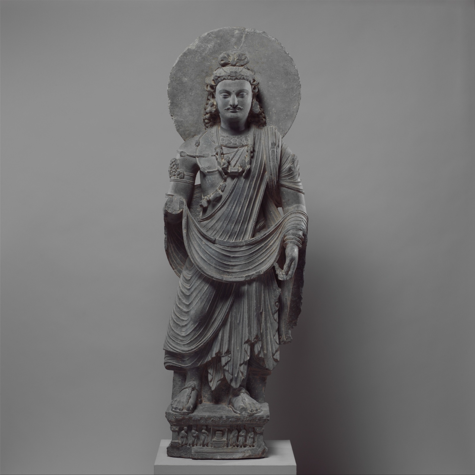 Statue de pierre grise d'un bodhisattva debout, auréolé, paré de bijoux, drapé dans une étoffe aux plis profonds, sur un socle sculpté