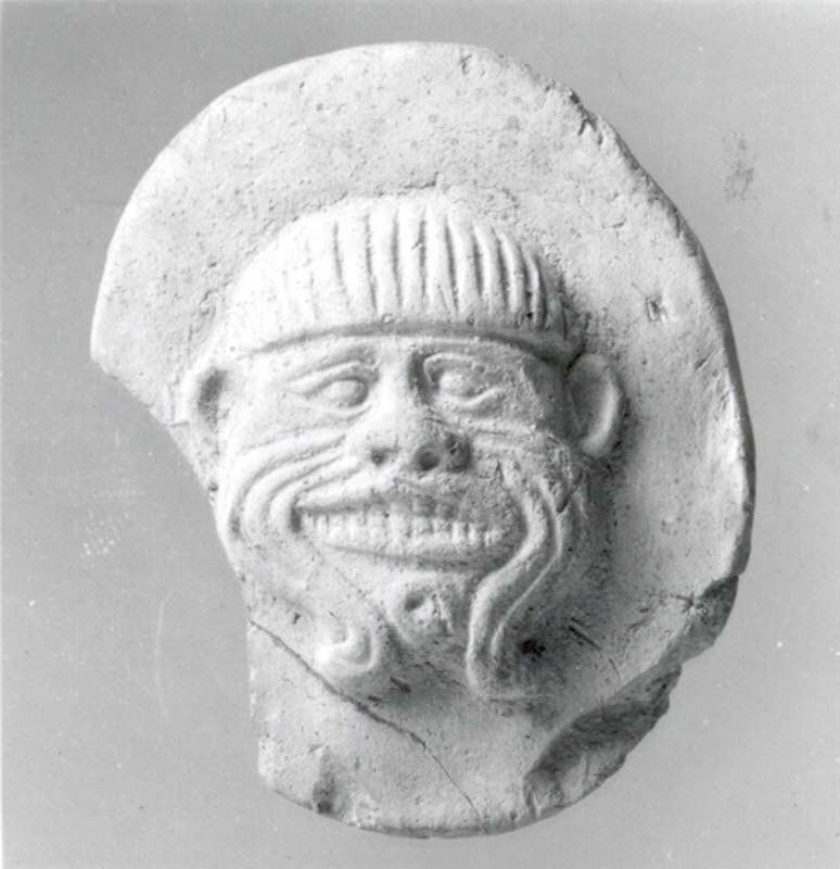 Petite plaque de terre cuite : un visage grimaçant aux traits creusés de sillons, coiffé d'un bonnet strié