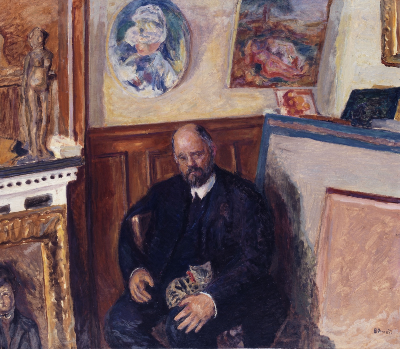 Un homme barbu en costume sombre, assis, un chat sur les genoux, entouré de tableaux accrochés ou posés contre le mur