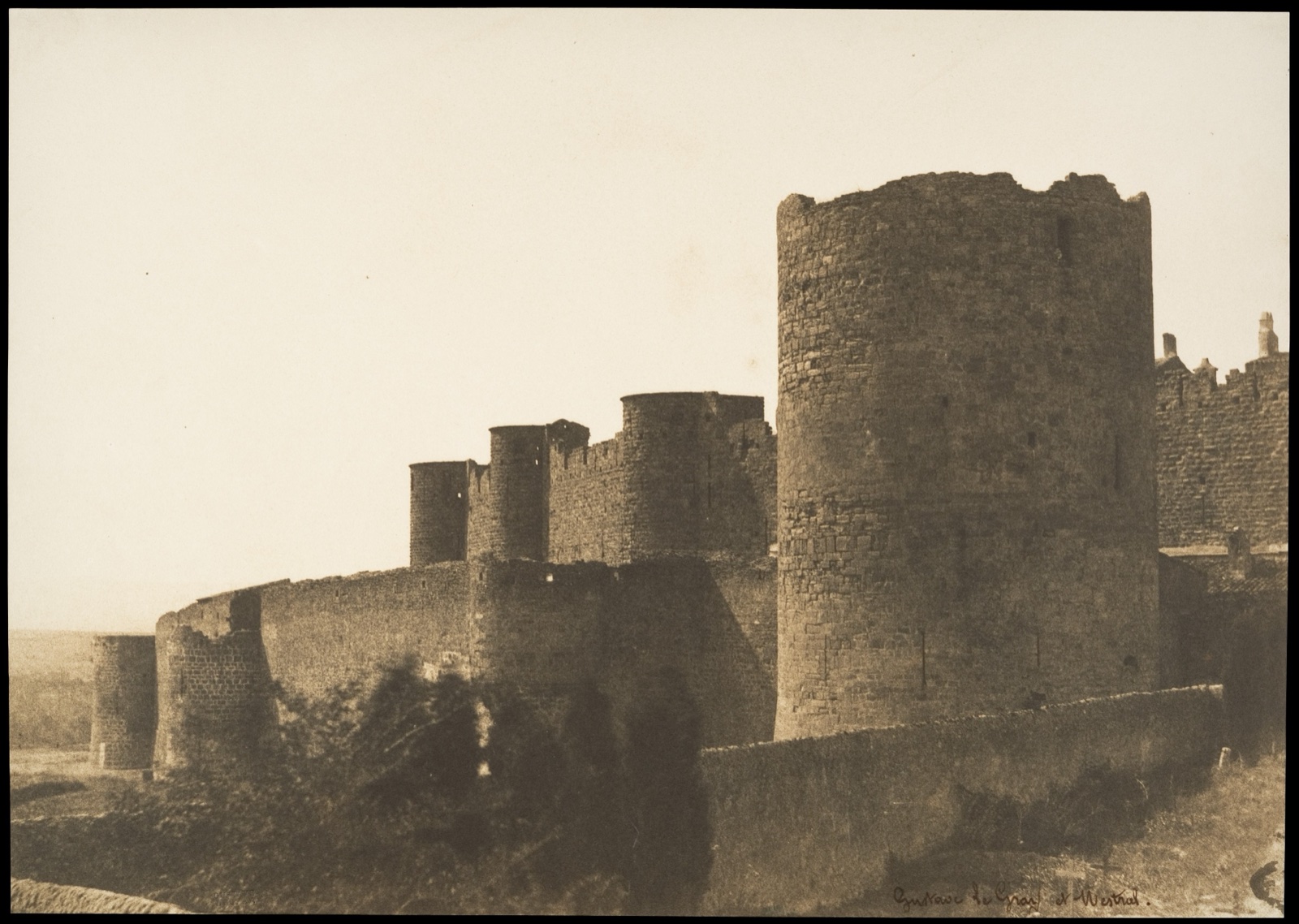 Photographie ancienne aux tons sépia : une longue muraille médiévale ponctuée de tours rondes, dont une grosse tour au premier plan, se détache sur un ciel pâle