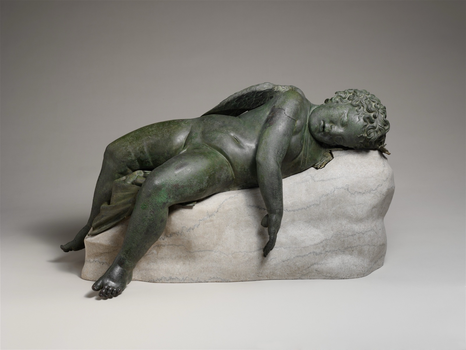 Statue de bronze vert sombre d'un jeune Éros ailé endormi, allongé sur le dos sur un rocher, un bras pendant