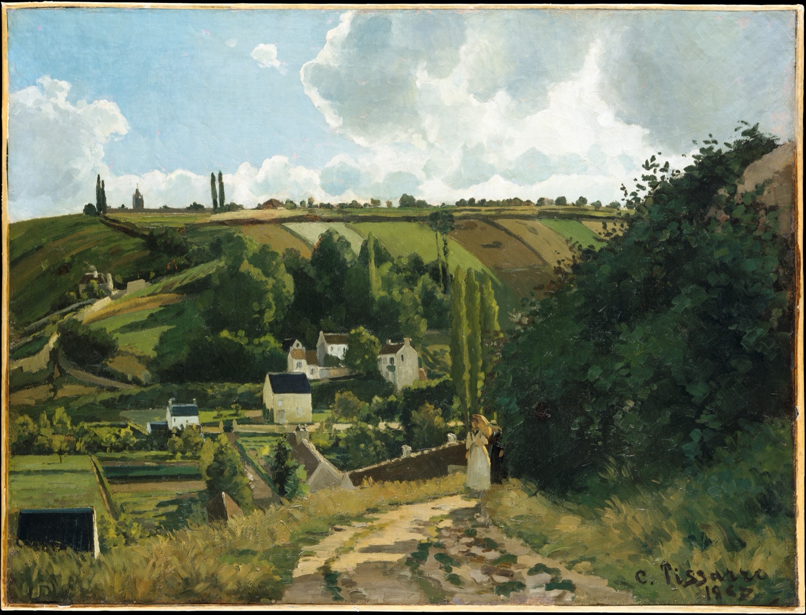 Un chemin de terre descend vers un village aux maisons blanches, entre des champs en pente et des bosquets, sous un ciel de nuages