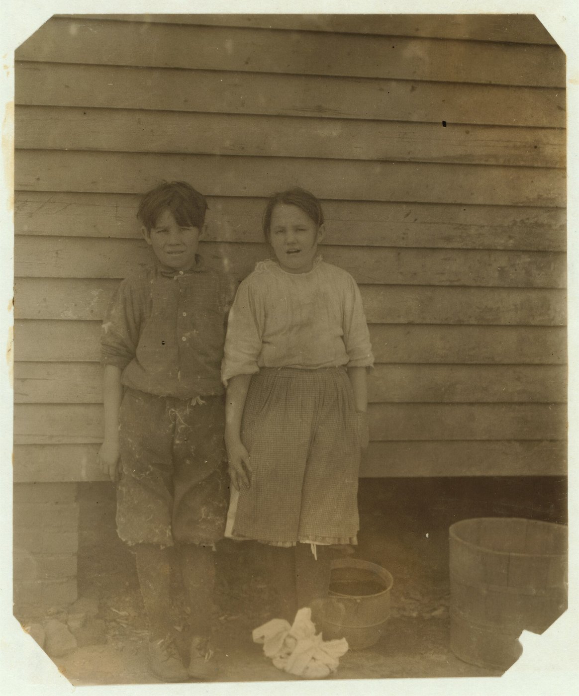Photographie ancienne : un garçon et une fille, pieds nus et vêtements tachés, debout côte à côte contre un mur de planches