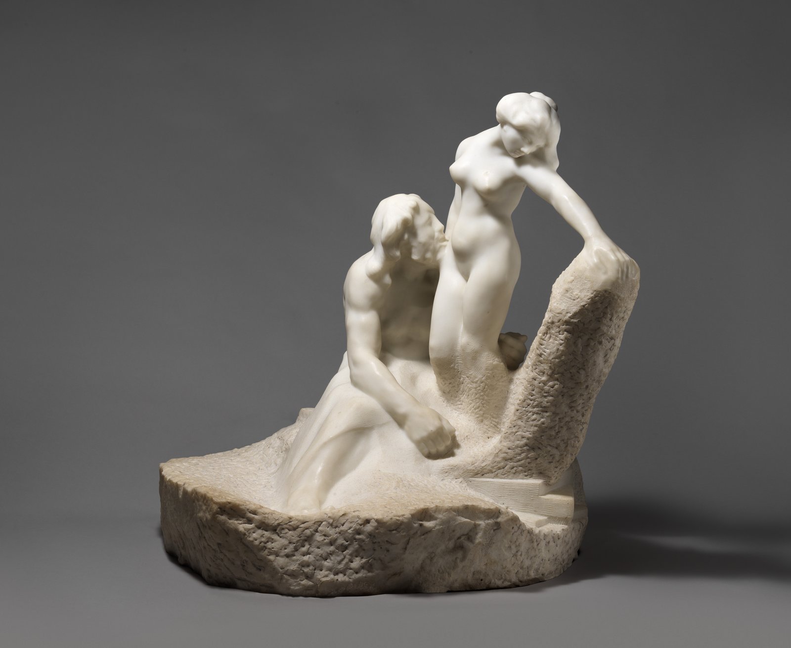 Groupe de marbre blanc : un homme assis contemple une jeune femme nue qui se dresse, les jambes encore prises dans le bloc de pierre brute
