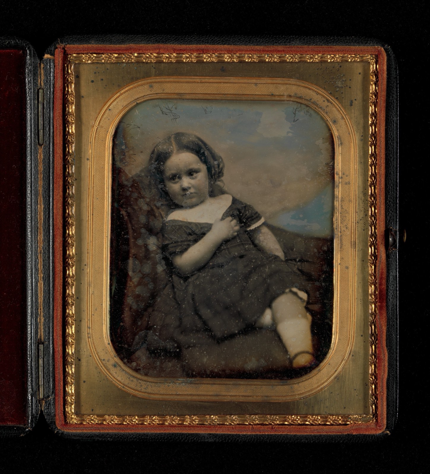 Petite photographie ancienne aux reflets argentés, dans un cadre doré : une fillette assise, en robe sombre, la main sur la poitrine